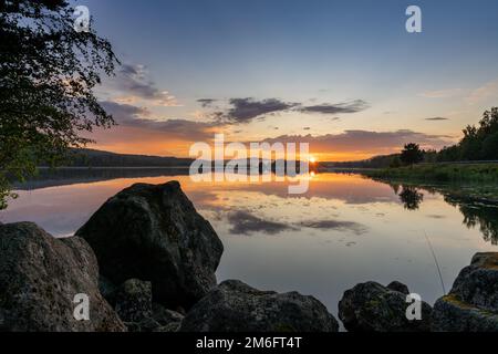 Tramonto colorato su un lago calmo con riflessi in acqua e rocce in primo piano Foto Stock