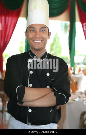 Uomo asiatico che indossa cappello e grembiule da chef sorride come lui  guarda le ciambelle sul tavolo mentre scrivi un notebook mentre si è seduti  in cucina Foto stock - Alamy