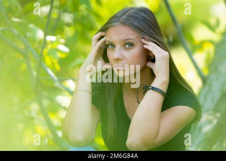 Una ragazza triste si siede su uno sfondo di fogliame, tiene la testa con le mani e guarda tristemente nel telaio Foto Stock