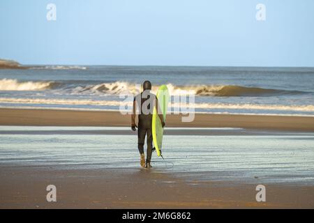 Vista posteriore di un surfista maschio che indossa una muta nera con una tavola da surf verde che cammina verso il mare a Cayton Bay, North Yorkshire, UK Foto Stock