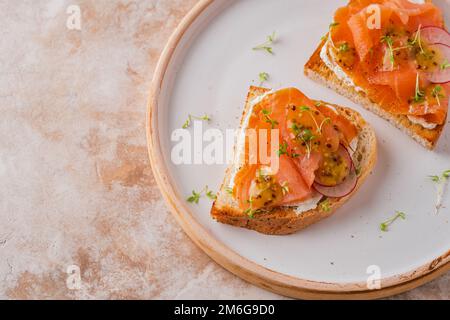 Due panini aperti, pane tostato con salmone, formaggio spalmabile, avocado, fettine di cetriolo su un tavolo di cemento bianco. Mattina sana colazione con pesce e tazza di d Foto Stock