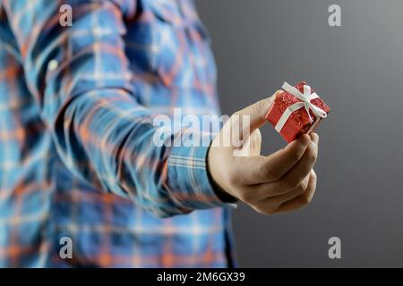 Una piccola scatola rossa con un regalo in mano di un uomo in una camicia blu a quadri. Piccola sorpresa. Concentrati sulla mano. Sfocatura regalo Foto Stock