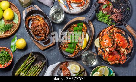 Set table con piatti di pesce - granchi cotti, gamberetti tigre, polpi alla griglia e calamari su padelle e piatti alla griglia in ghisa, vino bianco. Vista dall'alto. Foto Stock