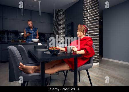 Aggiunta di gadget. Giovane famiglia che tiene e utilizza diversi dispositivi elettronici mentre si prende la colazione Foto Stock