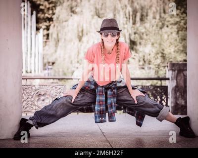 Una ragazza sorridente con pigtail e abiti casual stand con le sue gambe spalmata tra le colonne del parco. Ballerino di strada. Foto Stock