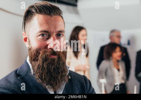 Primo piano ritratto di un giovane uomo d'affari hipster in ufficio con i colleghi. Viso sorridente con baffi pieni e barba lunga Foto Stock
