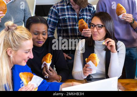 Un gruppo multietnico di colleghi di colleghi durante la pausa pranzo mentre mangiano croissant e dolci. Atmosfera festosa di risate e gioia Foto Stock