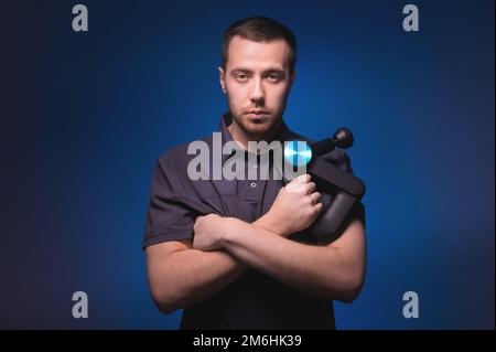 Ritratto di un massaggiatore professionista sicuro con un massaggiatore a percussione nelle sue mani. Tasto basso, sfondo blu. Onda d'urto massa Foto Stock