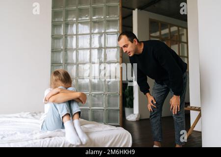 Ritratto di padre arrabbiato scoldendo, alzando la voce, urlando a triste testardo bambino bambino difficile seduto sul letto che abbraccia ginocchio, piangendo Foto Stock