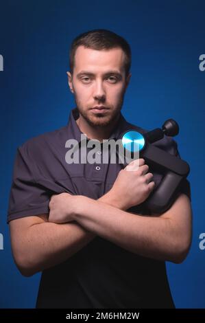 Ritratto di un massaggiatore professionista sicuro con un massaggiatore a percussione nelle sue mani. Tasto basso, sfondo blu. Onda d'urto massa Foto Stock