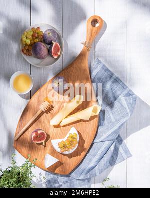 Formaggi assortiti con fichi, uva e miele su tavola di legno su fondo bianco con erbe fresche. Spuntini sani e deliziosi al mattino per br Foto Stock