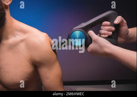 Primo piano massaggiatore a percussione nelle mani di un massaggiatore fa un massaggio delle spalle e della parte superiore della schiena a un cliente maschio sdraiato fare Foto Stock