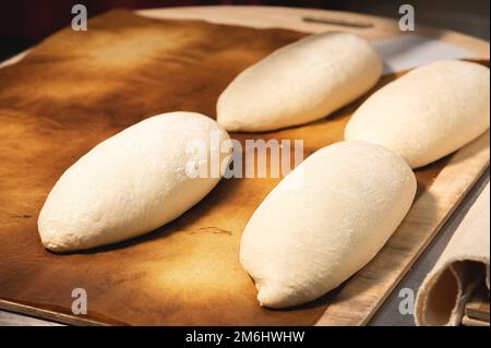 Su un asse di legno si trovano diverse focacce di pane francese crudo. Preparazione prima della cottura del pane artigianale Foto Stock