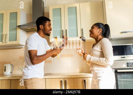 coppia ispanica cantando insieme tenendo un mestolo come un microfono in cucina Foto Stock