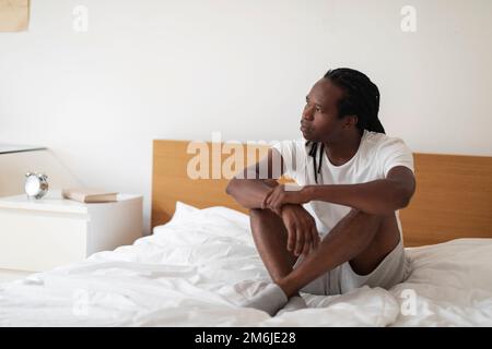 Ritratto di giovane maschio nero pensivo seduto sul letto e guardando lontano, Foto Stock