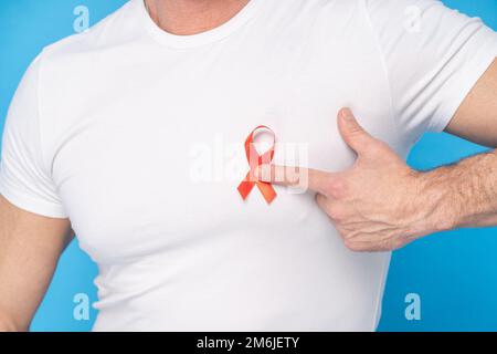 Uomo tenere rosso fiocco AIDS simbolo di consapevolezza su un cuore che indossa una t-shirt bianca isolato su uno sfondo blu. Medicina moderna Foto Stock