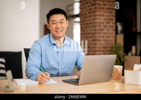 Felice uomo asiatico di mezza età che lavora su un notebook e prende appunti in notebook, sorridendo alla fotocamera, spazio libero Foto Stock