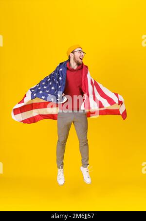 Felice, divertente giovane uomo in felpa con cappuccio rosso e bandiera USA jumping hight isolato su sfondo giallo. la libertà è nella tua vita. Allegro Foto Stock