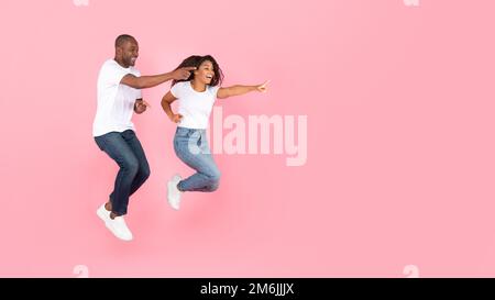 Uomo e donna neri eccitati che saltano in aria e puntano allo spazio libero, sfondo rosa studio, panorama Foto Stock
