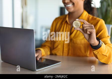 Commercio di criptovaluta. Donna d'affari nera irriconoscibile seduta alla scrivania con il laptop e che mostra il bitcoin dorato Foto Stock