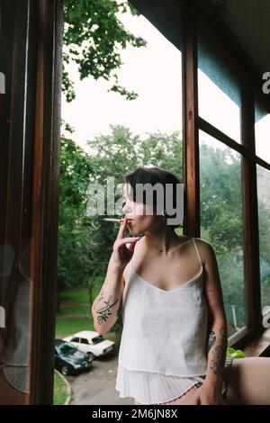 Donna con tatuaggi che fuma in piedi sul balcone Foto Stock