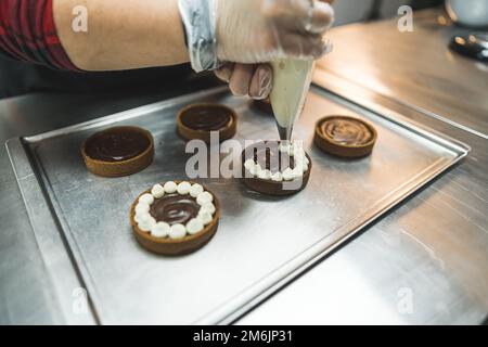 Le crostate poste su una teglia da forno essendo decorate con crema bianca da una persona che usa un sacchetto di pasticceria. Un panettiere che indossa un guanto trasparente. Sfondo sfocato. Foto di alta qualità Foto Stock