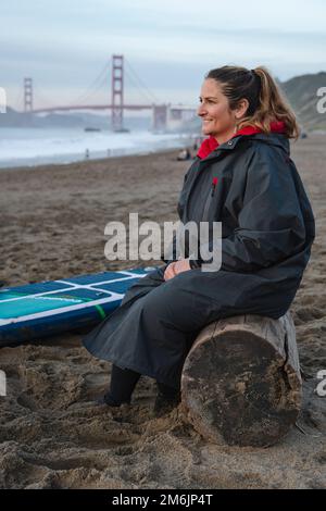 Donna sorridente seduta sul log da paddleboard alla spiaggia durante il tramonto Foto Stock