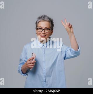 Gorgeous haired gorgeous maturo businesswoman con lo smartphone in mano che osserva la macchina fotografica che lavora o che acquista in linea o che controlla così Foto Stock