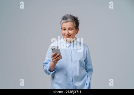 Splendida donna con capelli grigi maturi tenere in mano smartphone guardando la fotocamera di lavoro o di shopping online o di controllo sul social med Foto Stock