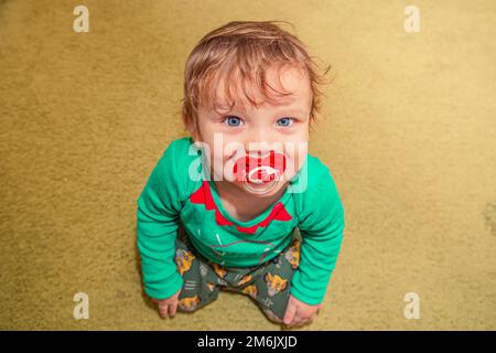 Allegro ragazzo sorridente dai capelli rossi siede sul pavimento con un succhietto in bocca Foto Stock