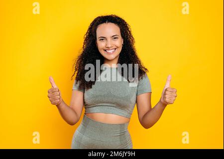 Foto di felice bella donna sportiva brasiliana o ispanica con capelli ricci in abbigliamento sportivo, con gesto pollice su, guarda la macchina fotografica, sorridendo, in piedi su sfondo isolato arancione Foto Stock