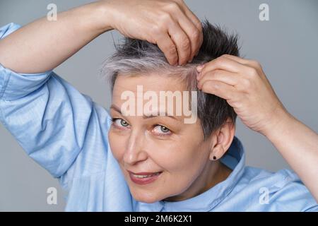 Donna graziosa in 50s che controlla la sua pelle dei capelli o del cranio nello specchio. Bella donna dai capelli grigi che si occupa di dandruff problema check in Foto Stock