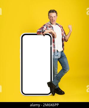 Puntando su un enorme e gigantesco smartphone con schermo bianco giovane uomo appoggiato su di esso gesturing con un'altra mano vestita in casual isolat Foto Stock