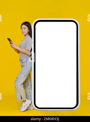 Ragazza asiatica sorridente con telefono in mano appoggiato su enorme smartphone gigante, tablet digitale con schermo bianco, nero in piedi w Foto Stock