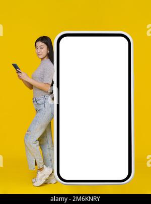 Ragazza asiatica sorridente con telefono in mano appoggiato su enorme smartphone gigante, tablet digitale con schermo bianco, nero in piedi w Foto Stock