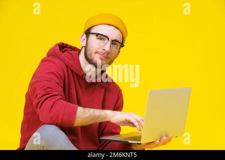 Giovane uomo hipster seduto sul pavimento con laptop in felpa con cappuccio rossa che studia o lavora. Freelance intelligente uomo caucasico con lap Foto Stock