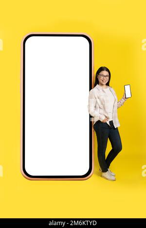 Affascinante donna asiatica di mezza età con telefono in mano che mostra lo schermo bianco appoggiato su gigantesco, enorme smartphone con carta bianca Foto Stock