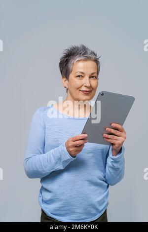 Donna dai capelli grigi con PC tablet in mano che lavora online. Bella donna in 50s indossare blusa blu isolato sul grigio. Peopl maturo Foto Stock