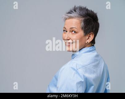 Capelli grigi donna matura posa in piedi indietro mezzo giro guardando sopra la spalla alla macchina fotografica e sorridere indossando camicia blu isolato su w Foto Stock