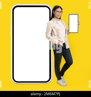 Mostrando schermo bianco sul telefono affascinante donna asiatica di mezza età appoggiata indietro su gigantesco, enorme smartphone con schermo bianco che indossa c Foto Stock
