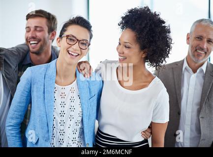 Un altro buon lavoro. Ritratto di due colleghi che sorridono in ufficio con colleghi sullo sfondo. Foto Stock