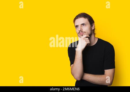 Bell'uomo caucasico che pensa con il mento che tocca la mano isolato su sfondo giallo studio con spazio copia. Concetto di stile di vita della gente. Soluzione Foto Stock