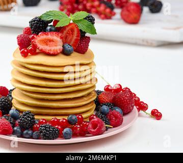 Accatastate le frittelle cotte con la frutta in un piatto rotondo su un tavolo bianco Foto Stock