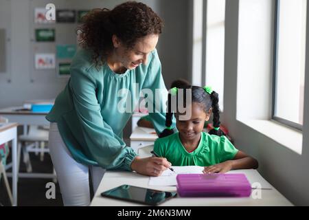 Giovane insegnante caucasica che assiste la scolarista afroamericana elementare scrivendo sul libro Foto Stock