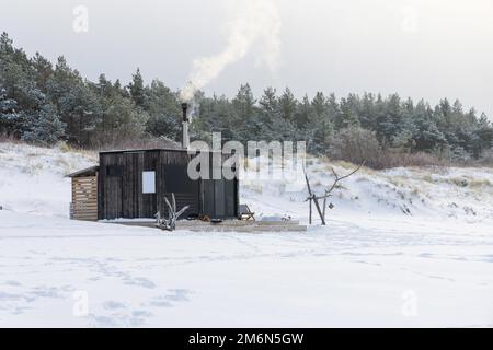 Sauna all'aperto in legno con fumo che esce dal camino in una bella giornata invernale fredda e innevata al Mar Baltico. Benessere e stile di vita sano. Foto Stock