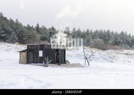 Sauna all'aperto in legno con fumo che esce dal camino in una bella giornata invernale fredda e innevata al Mar Baltico. Benessere e stile di vita sano. Foto Stock