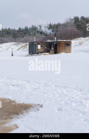 Sauna all'aperto in legno con fumo che esce dal camino in una bella giornata invernale fredda e innevata al Mar Baltico. Benessere e stile di vita sano. Foto Stock