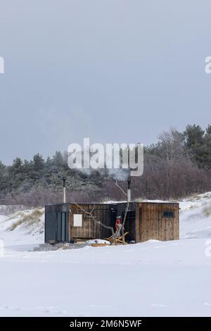 Sauna all'aperto in legno con fumo che esce dal camino in una bella giornata invernale fredda e innevata al Mar Baltico. Benessere e stile di vita sano. Foto Stock