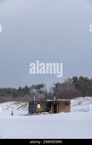 Sauna all'aperto in legno con fumo che esce dal camino in una bella giornata invernale fredda e innevata al Mar Baltico. Benessere e stile di vita sano. Foto Stock