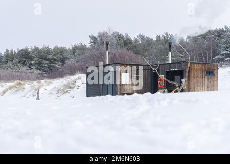 Sauna all'aperto in legno con fumo che esce dal camino in una bella giornata invernale fredda e innevata al Mar Baltico. Benessere e stile di vita sano. Foto Stock